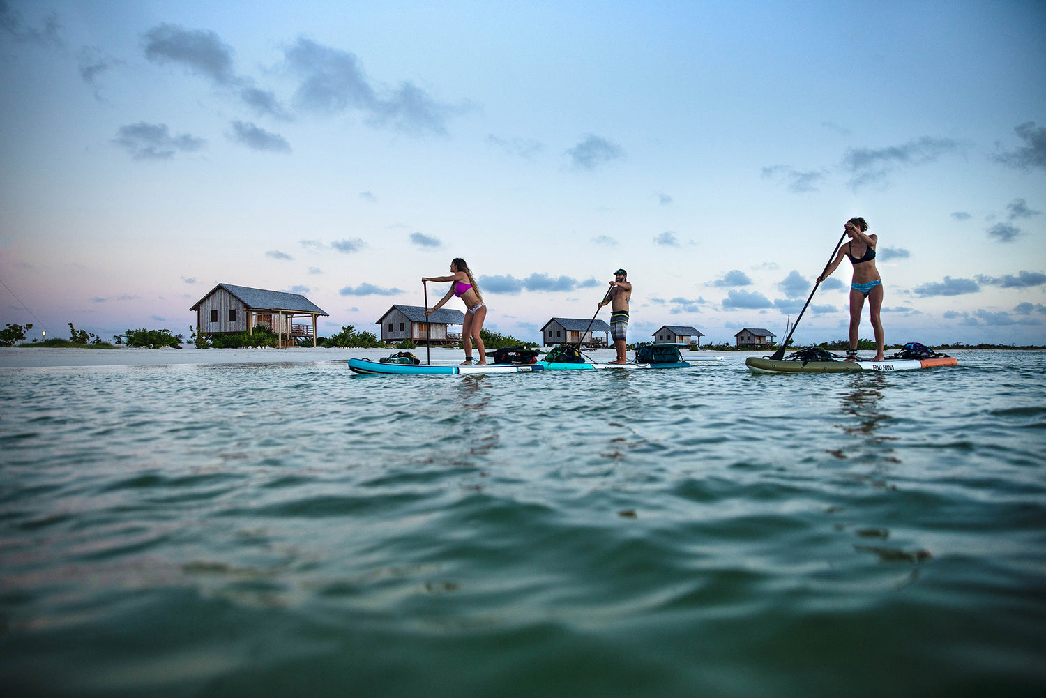 three paddleboards paddling on the ocean at dush with villas on the beach in the background