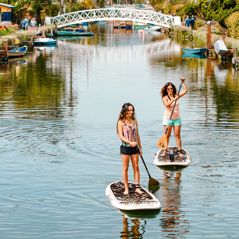two people paddling paddle boards on a canal having fun with a white bridge in the background