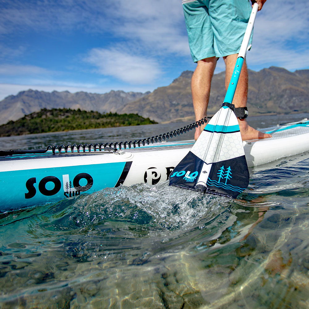 a close up of a paddle leaving the water next to a stand up paddle board on a lake with mountains in the background