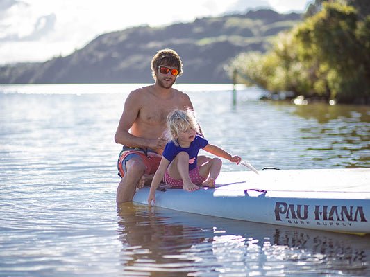 A man and child sat on a giant inflatable SUP on a lake with native new zealand bush in the background