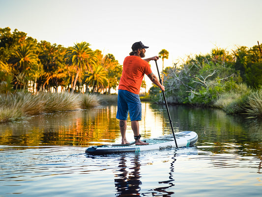 a man paddling an inflatable paddle board into the sunset between palm trees on a river