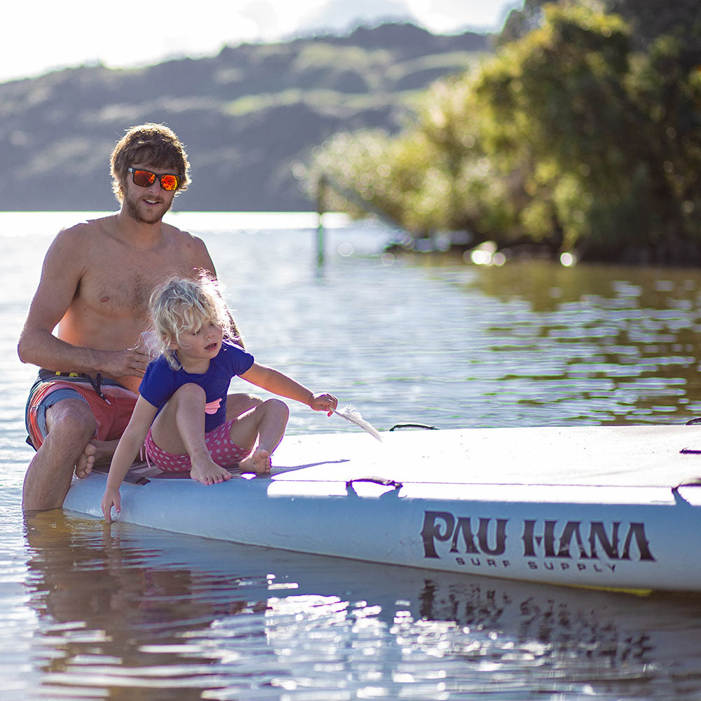 a man and child sitting on a giant inflatable paddle board on a lake with bushes and hills in the background