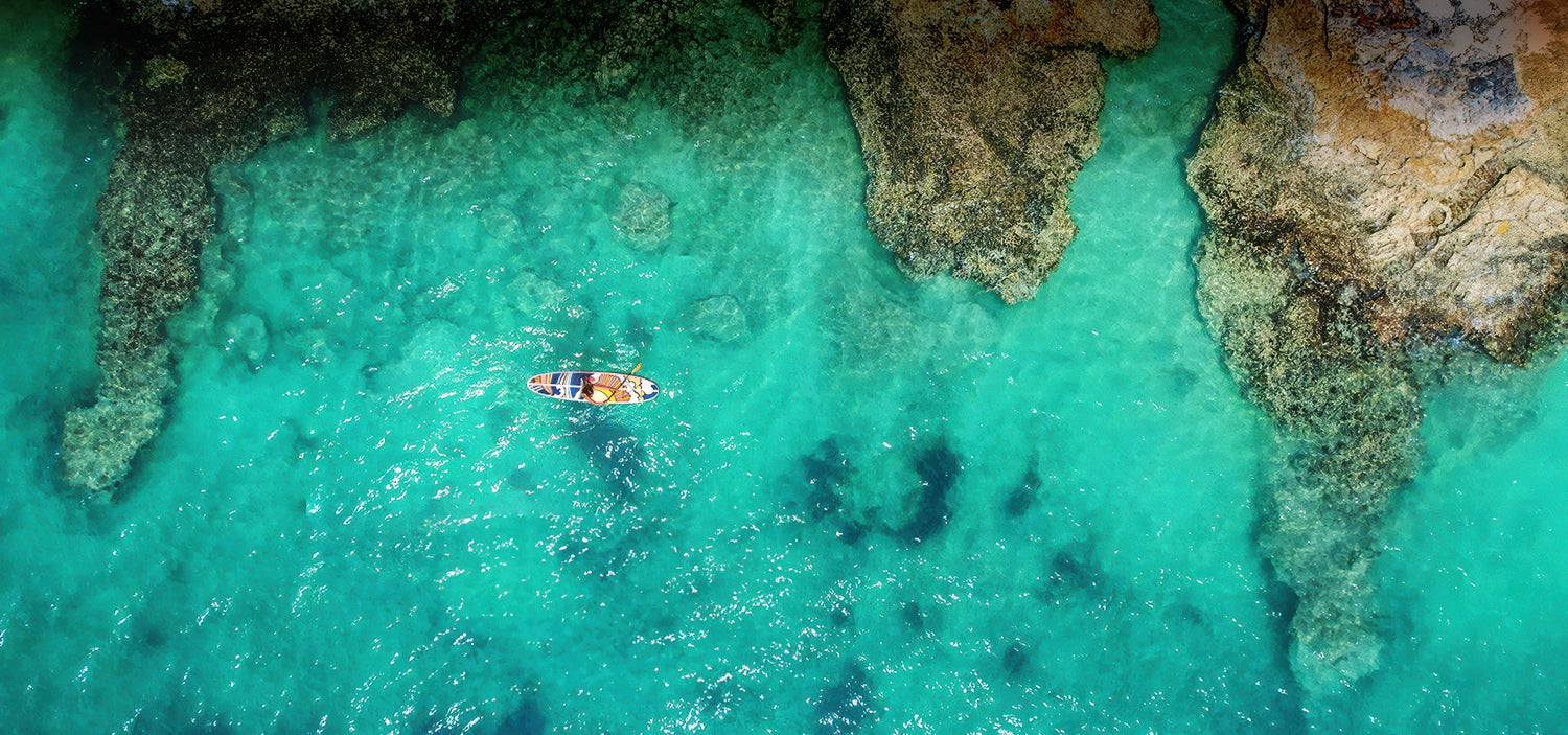 an aerial image of a colourful paddleboard on a bright green ocean with an outcrop of rocks