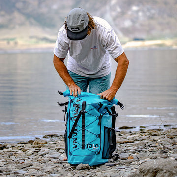 a man on a pebble beach packing up the solo sup backcountry paddleboard into the drybag backpack