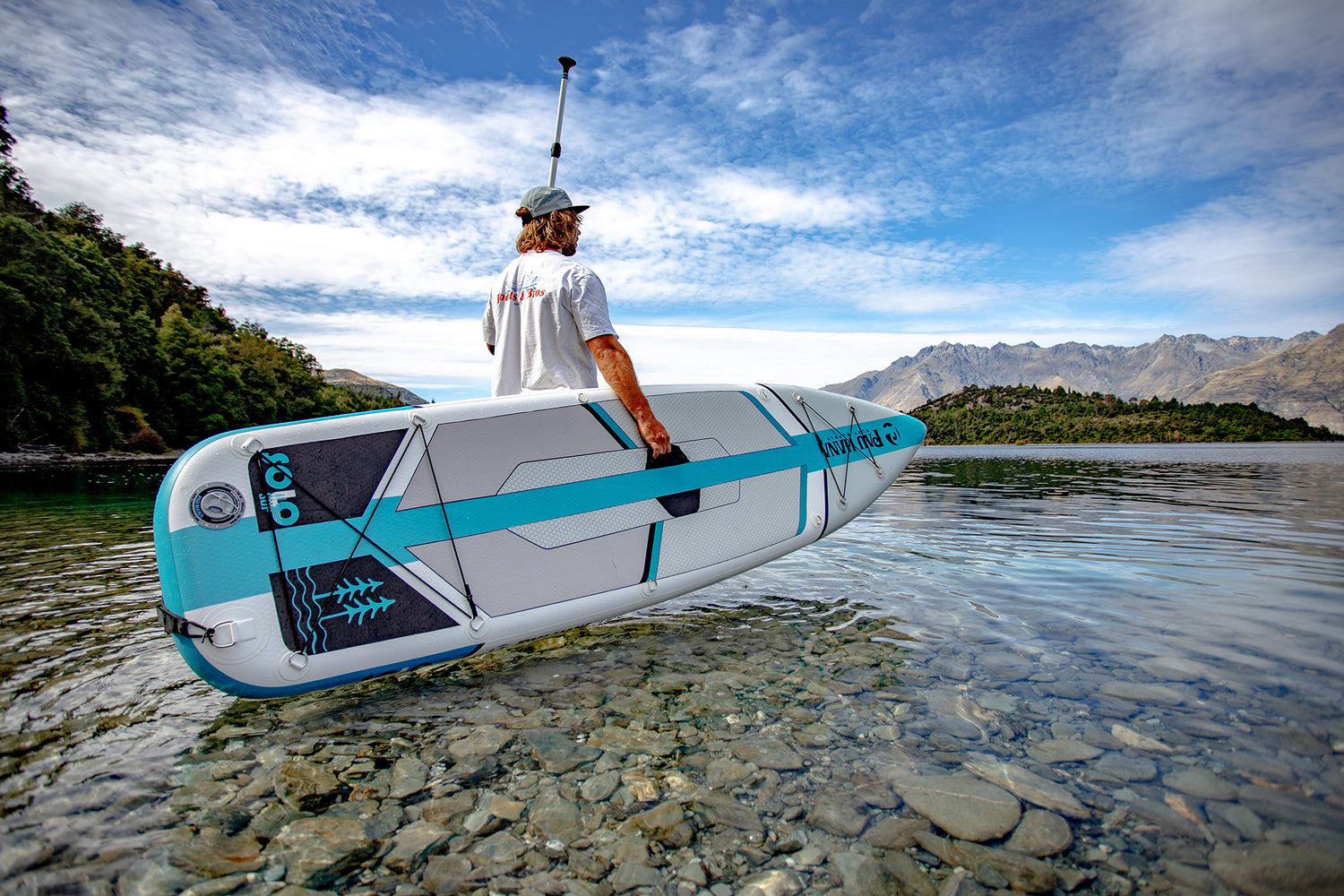 man standing in shallow water holding an inflatable paddleboard with mountains in the background