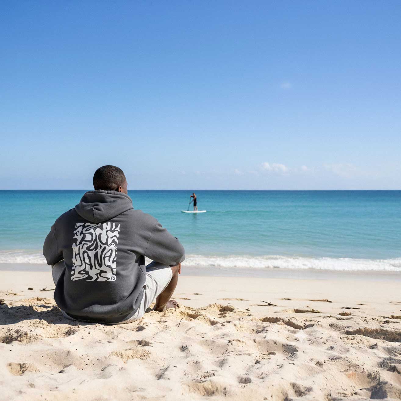 Person sitting on a beach looking at the ocean with another person paddleboarding.