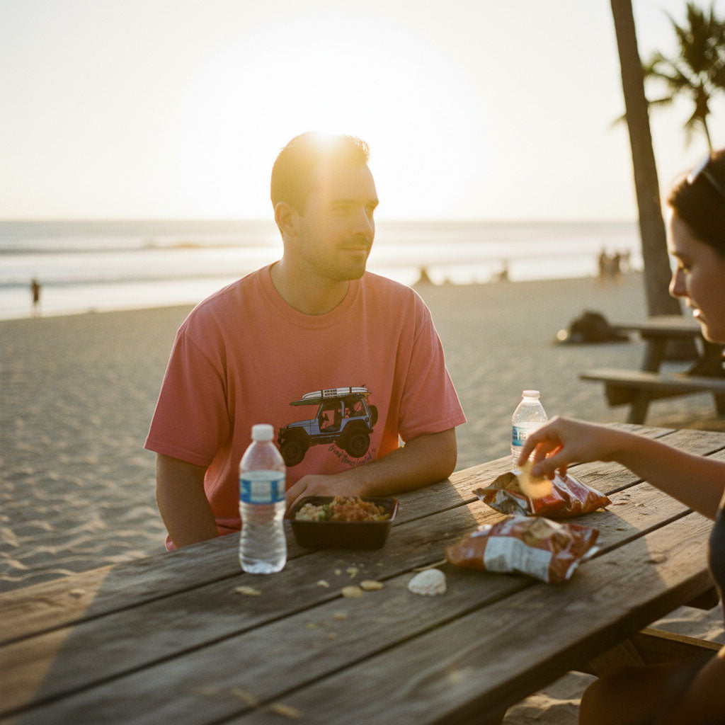 Two people sitting at a picnic table by the beach with food and drinks.