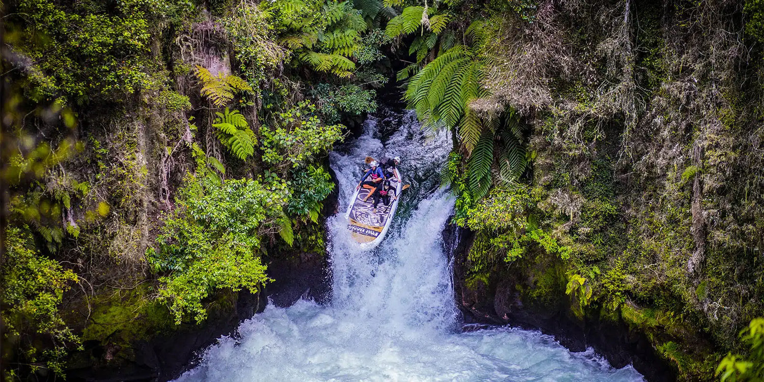 three people on a giant paddleboard going over a waterfall surrounded by native new ealand bush