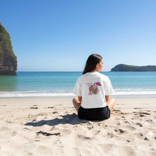 Person sitting on a beach wearing a white t-shirt with a design on the back, looking at the ocean.